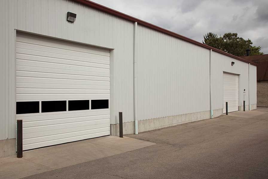 White industrial warehouse with roll-up doors along an asphalt lot under an overcast sky