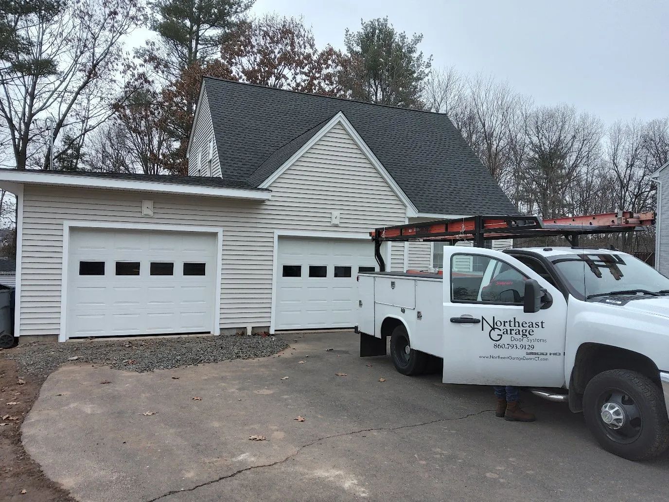 White service truck parked beside a two-car garage with a dark shingled roof and bare trees behind it
