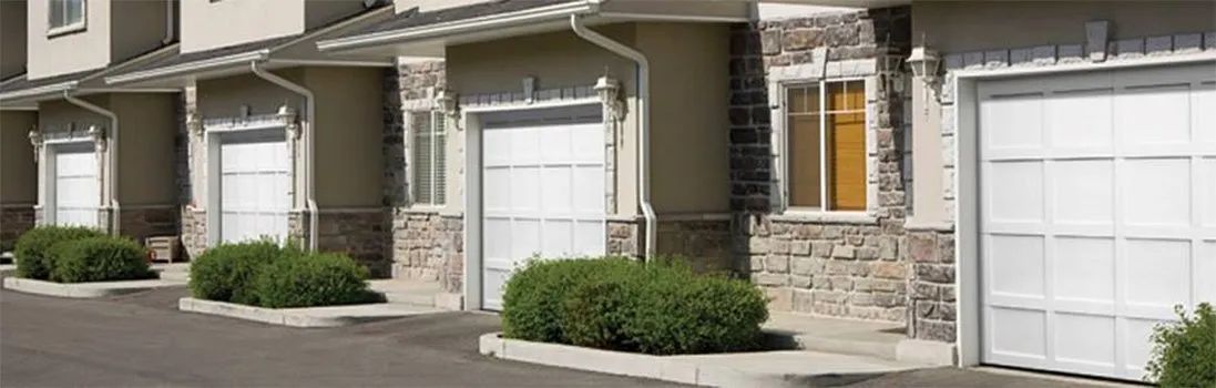 Row of beige townhouses with white garage doors and trimmed shrubs along a residential street