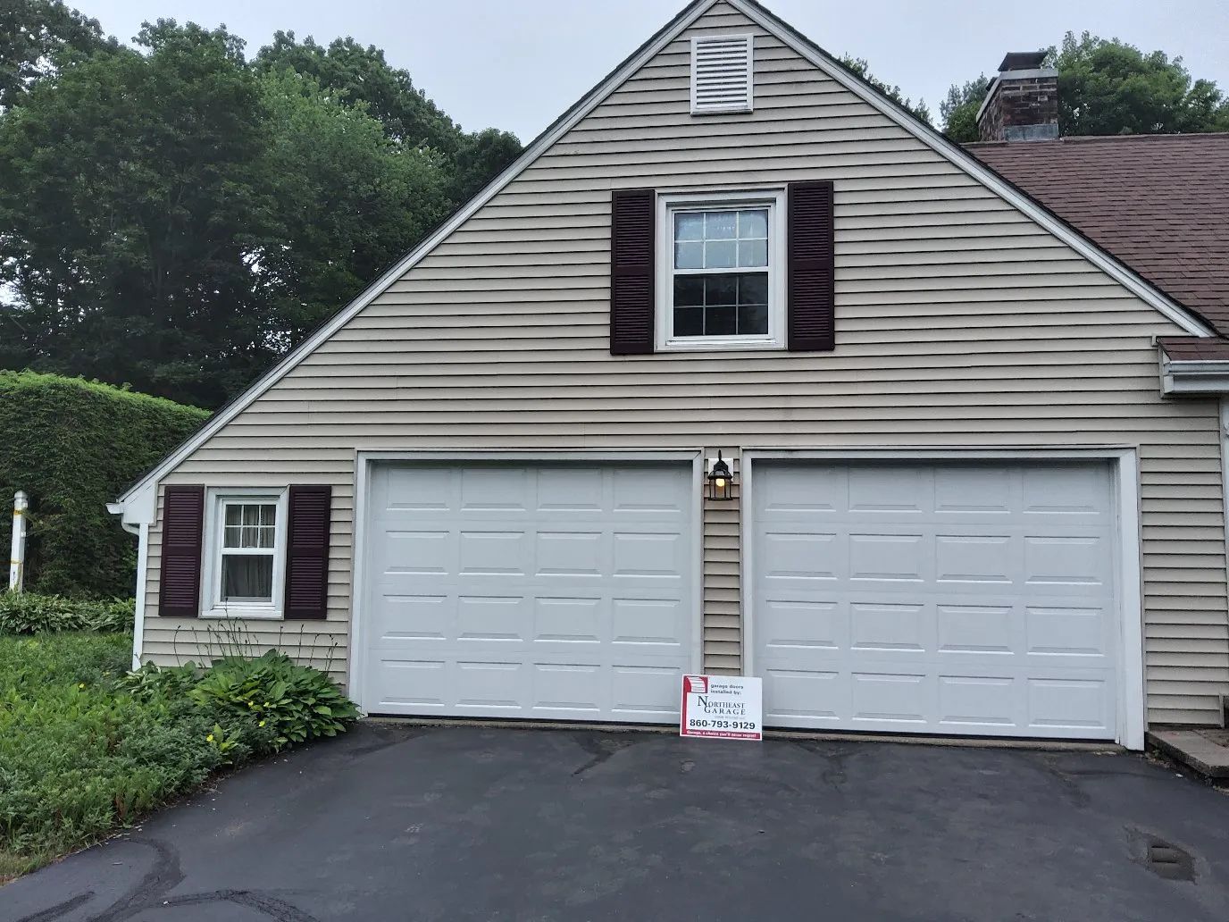 Suburban house with two white garage doors, tan siding, and a small sign at the driveway entrance