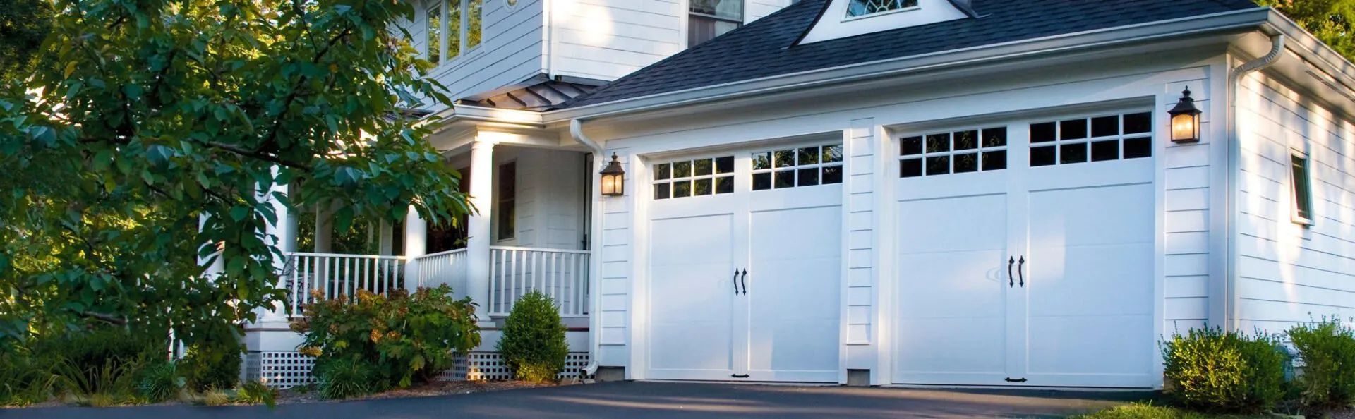 White garage doors on a sunlit suburban house with trees and a front porch