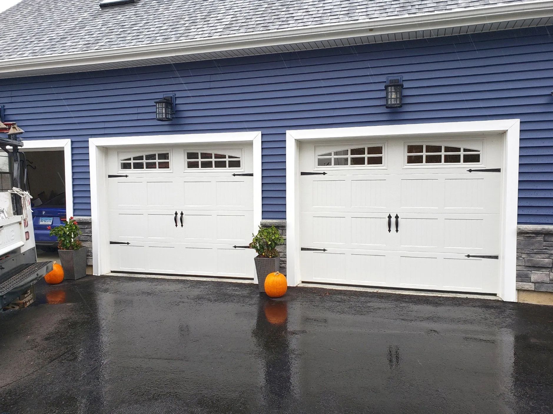 Two white garage doors on a blue house with wet driveway and orange pumpkins by the entrance