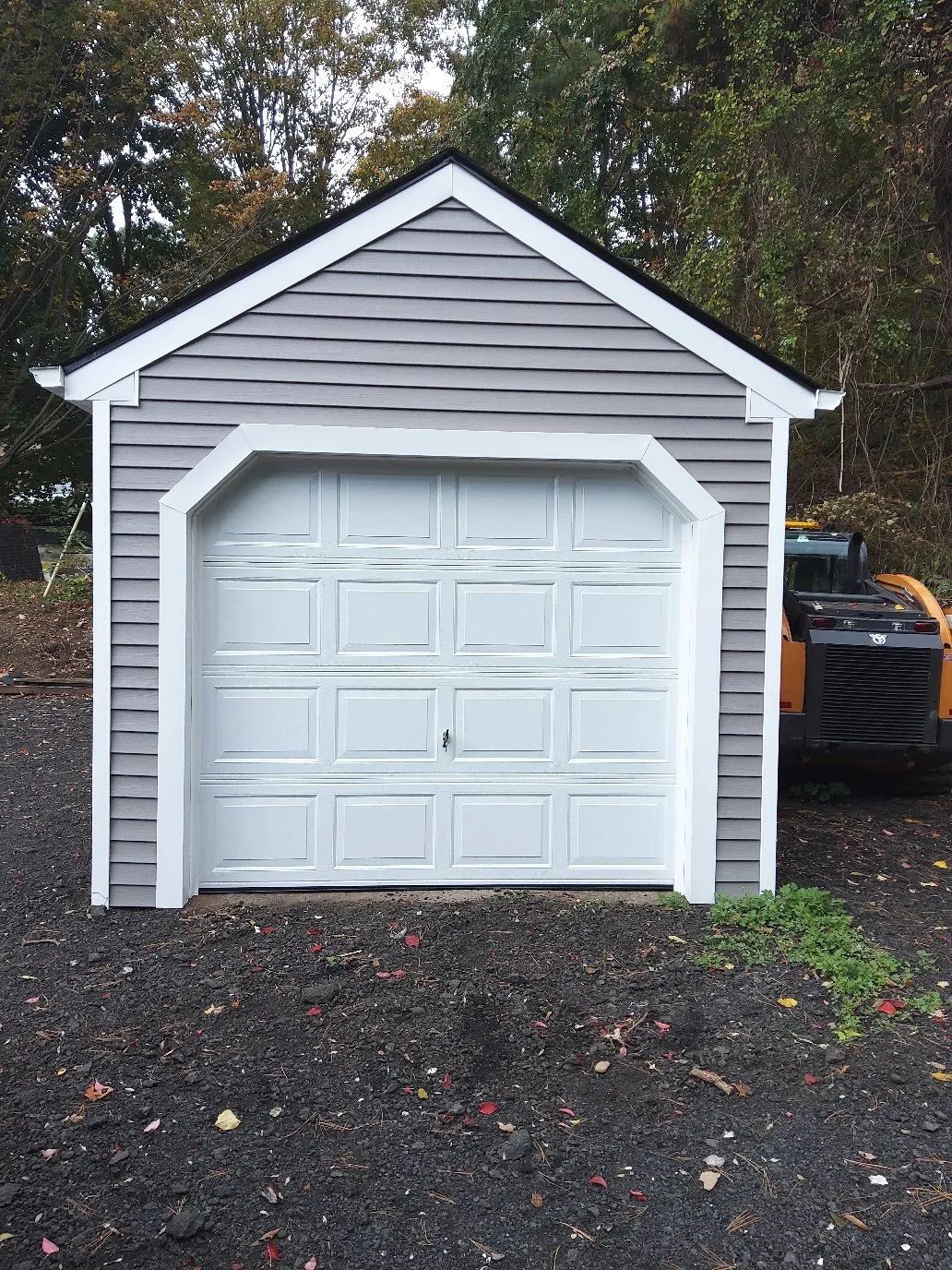 Small white detached garage with gray siding and a white door on a gravel driveway, trees behind.