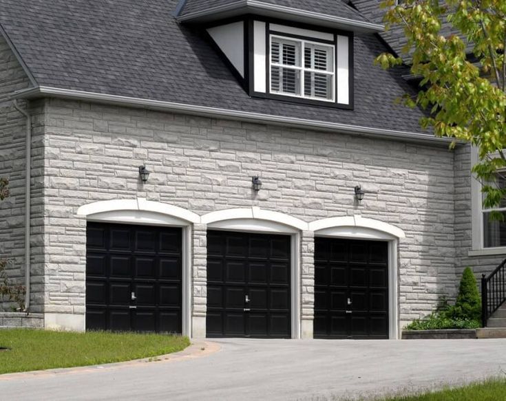 Three black garage doors on a gray stone house with a dark shingled roof.