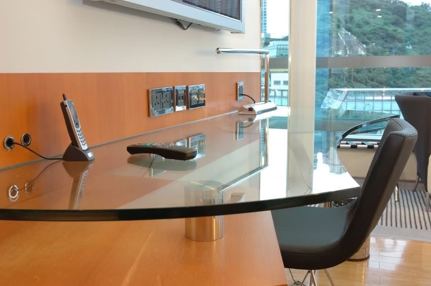 Glass desk with phone, remote, power outlets, and a view of buildings and nature through a window.