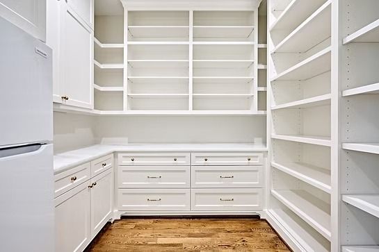 White pantry with built-in shelves, drawers, and cabinetry, featuring a refrigerator and wooden floor.