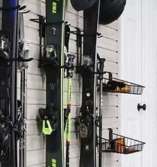 Skis and poles on a wall-mounted storage system with black baskets, on a light gray wall.