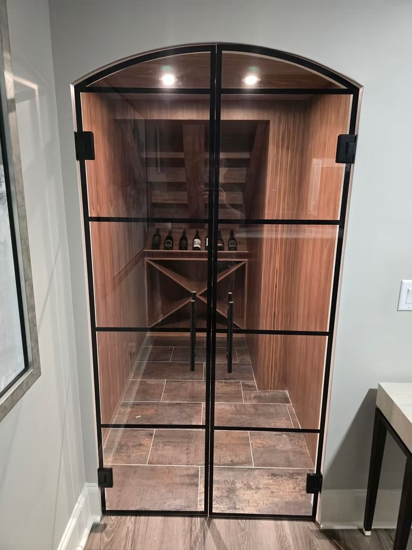 Glass-doored wine cellar with black metal frame; interior with wooden shelves and brick-like floor.