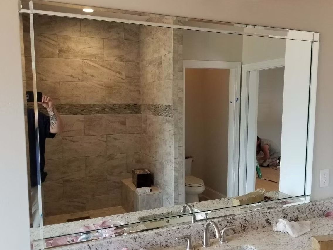 Bathroom mirror reflecting a shower, toilet, and person. Granite countertop with faucets. Beveled edges.
