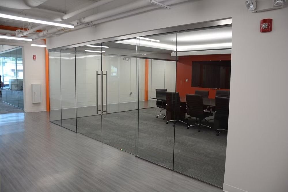Glass-walled conference room with table, chairs, and orange accent wall. Light grey floors and white ceiling.