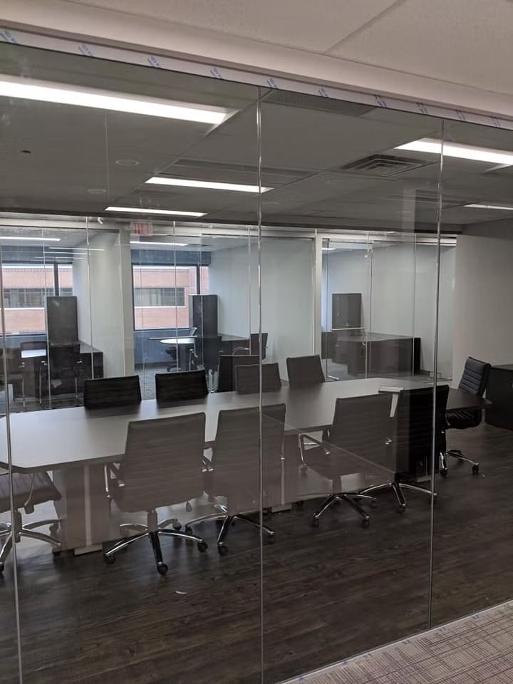 Conference room with long table, chairs, and glass walls. Buildings visible through the windows.