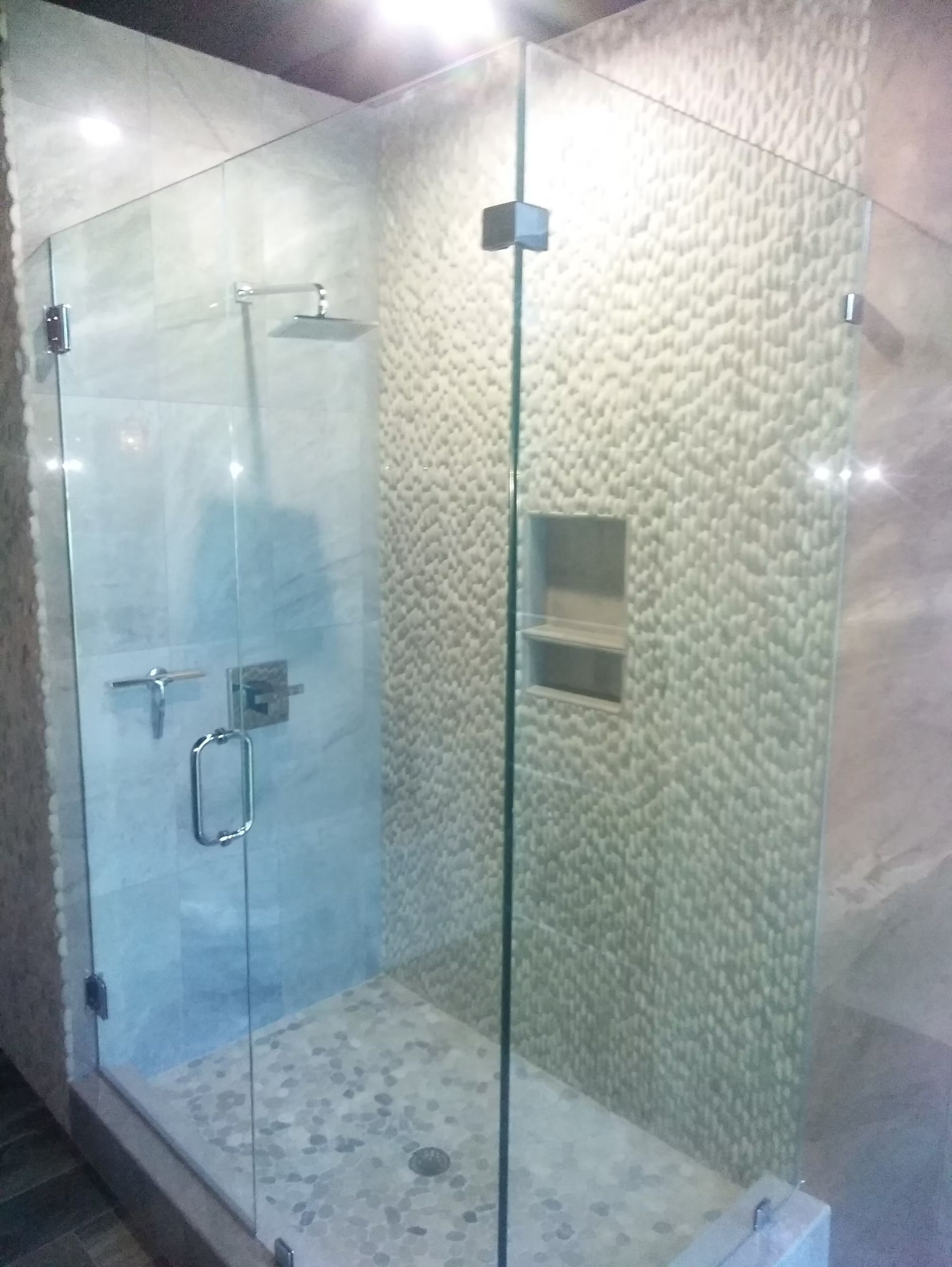 Glass-enclosed shower with pebble floor and textured beige tile walls. Stainless steel fixtures and a built-in shelf.