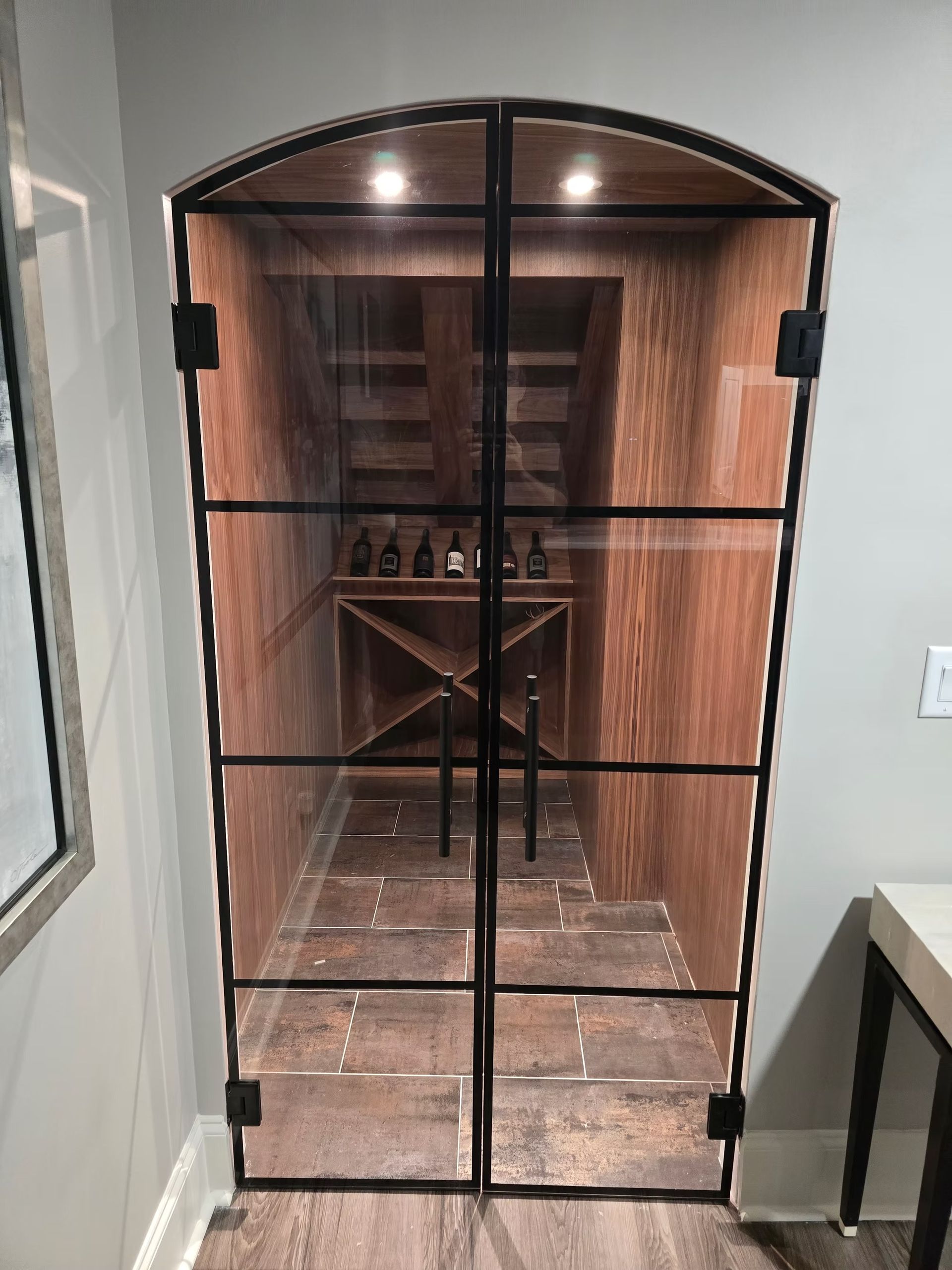 Wine cellar behind a black-framed glass door, wooden interior, with bottles visible inside, recessed into a wall.
