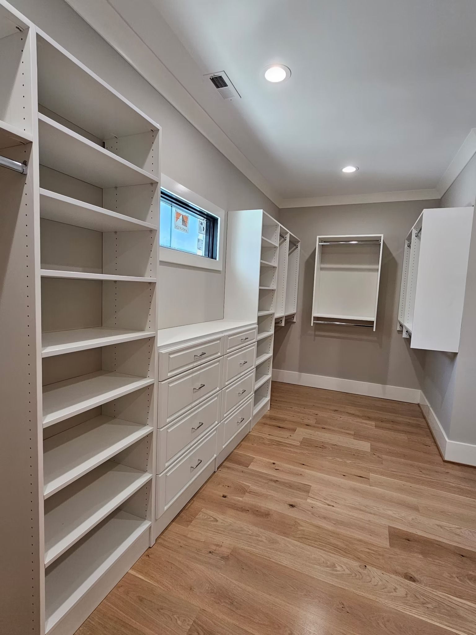 Walk-in closet with white shelving, drawers, and wooden floor. A small window is in the center.