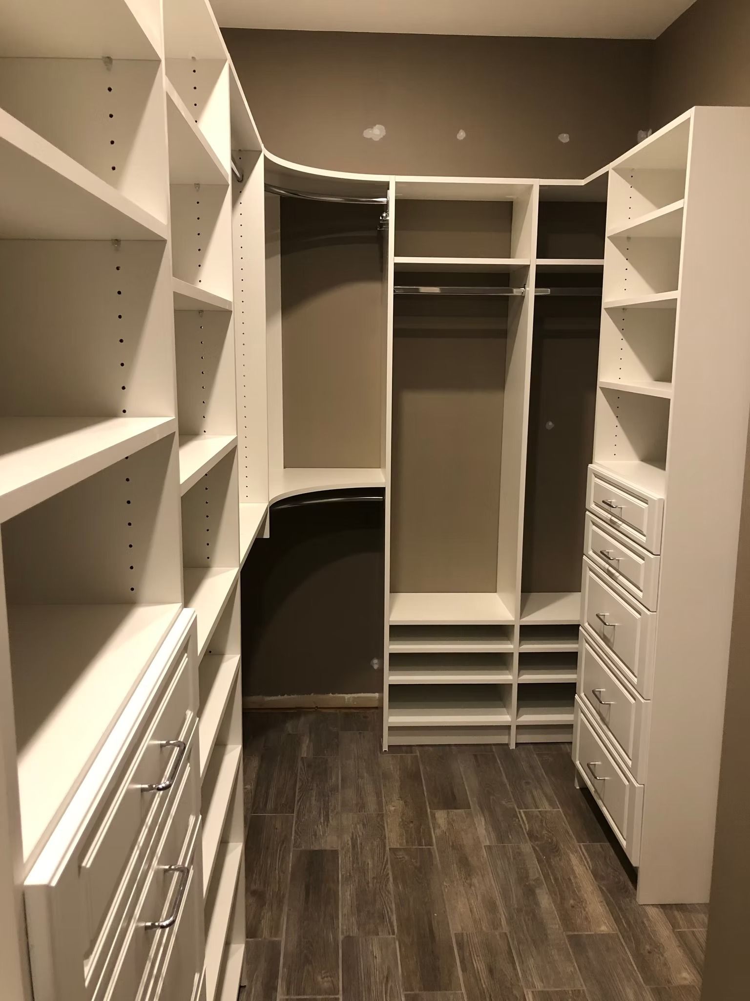 White closet organizer with shelves, drawers, and hanging rods; set in a walk-in closet with wood-look tile flooring.