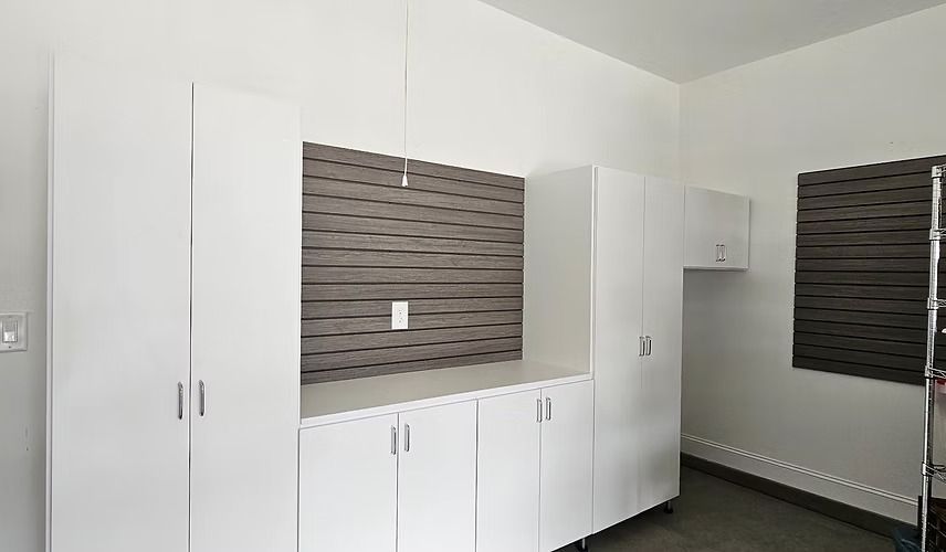 White storage cabinets in a garage against a gray wood-paneled wall and a window with blinds.