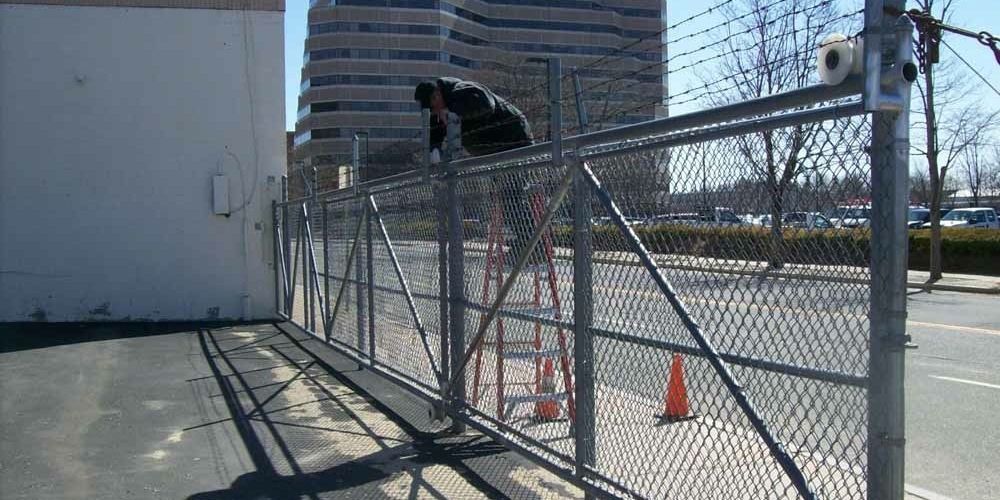 Person climbing a chain-link fence beside a sidewalk, with orange cones nearby and buildings in the background