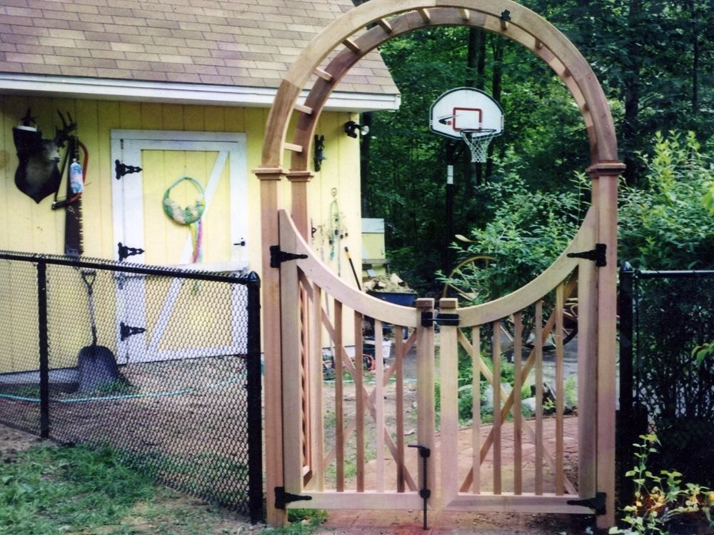 Wooden garden gate with circular arch framing a basketball hoop beside a yellow house and fenced yard