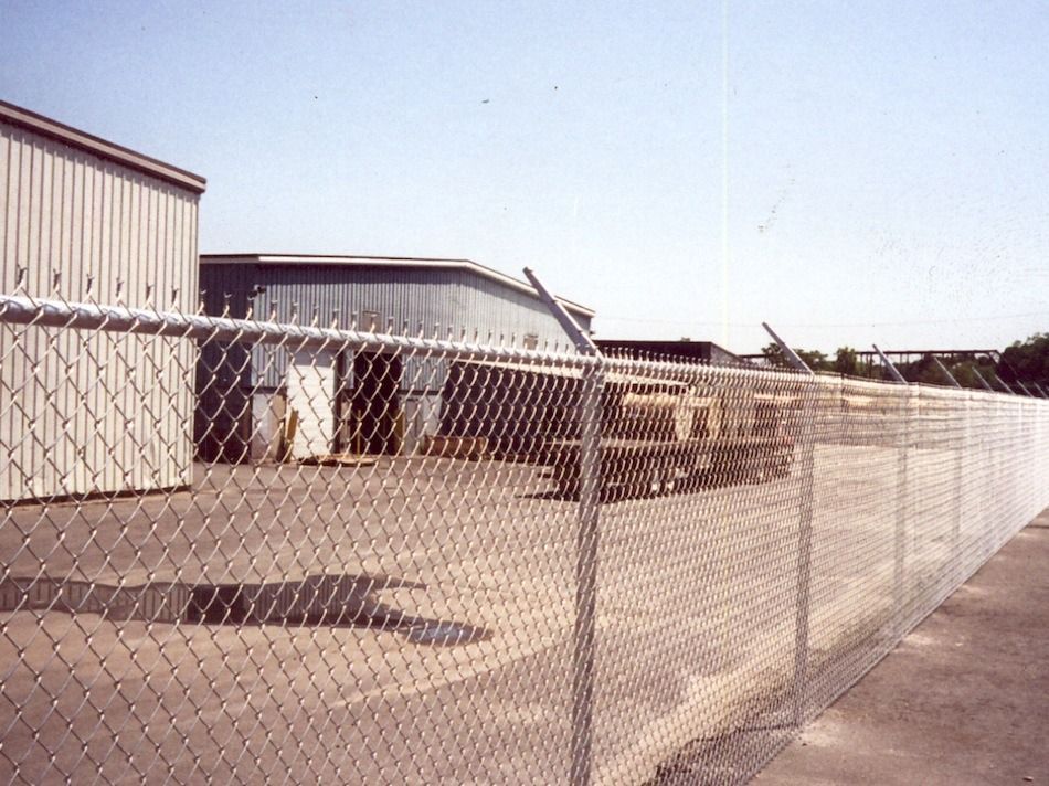 Chain-link fence along an empty industrial building and parking lot under a clear sky.