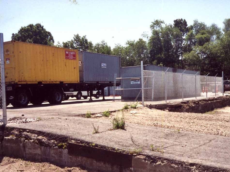 Yellow and gray freight trailers beside a chain-link fenced lot on a sunny street.