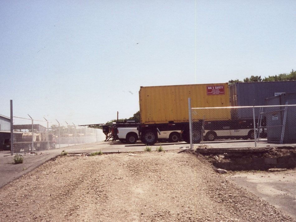 Yellow semi-truck blocking a gravel road near chain-link fencing in an industrial lot