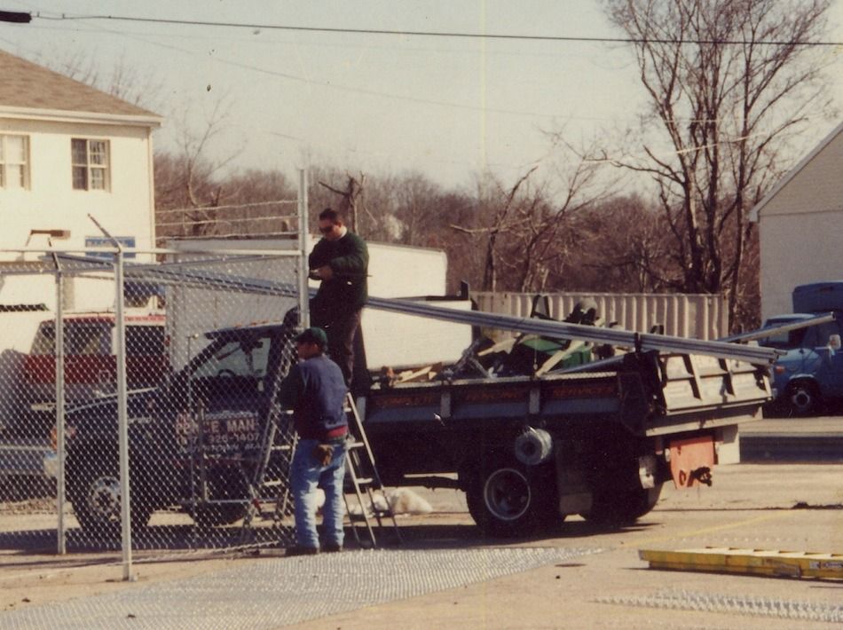 People standing by a black pickup truck at a chain-link gate in a residential yard.