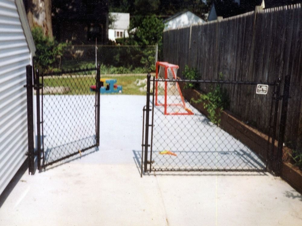 Open chain-link gate to a concrete backyard with a small red play structure and wooden fence
