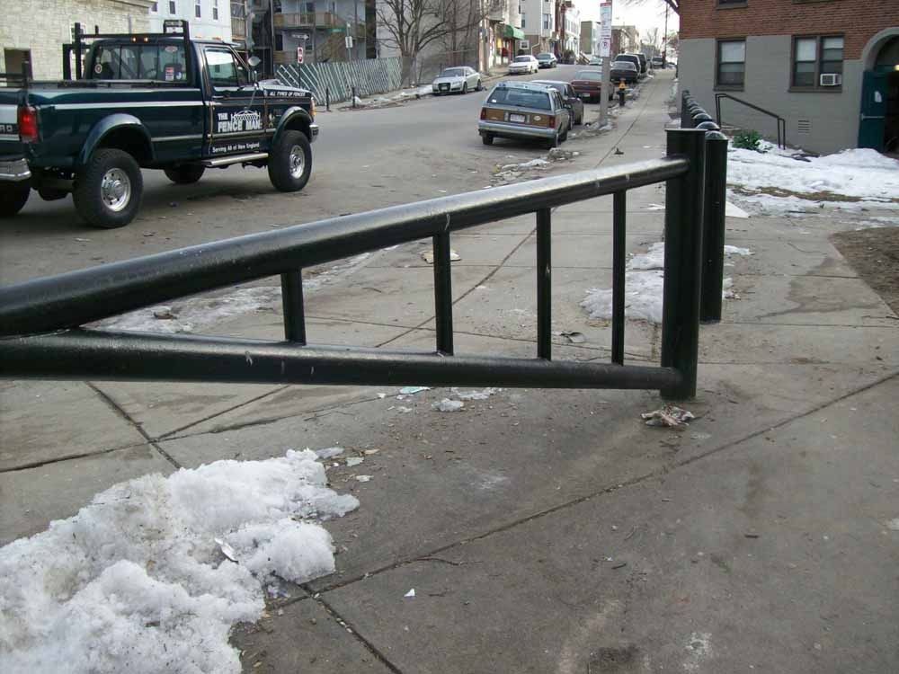 Broken metal gate blocking a snowy sidewalk beside a city street with parked cars