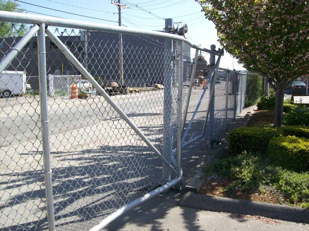 Chain-link gate beside a paved lot, with metal braces and bushes along the fence