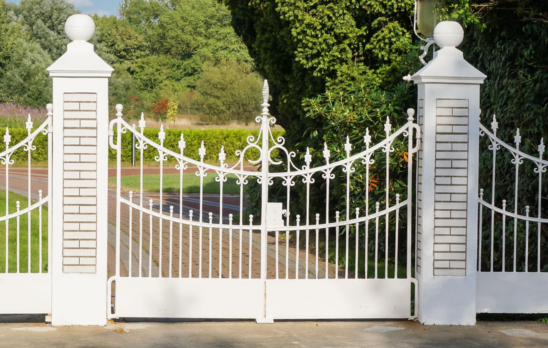White wrought-iron driveway gate with matching stone pillars in front of a green garden