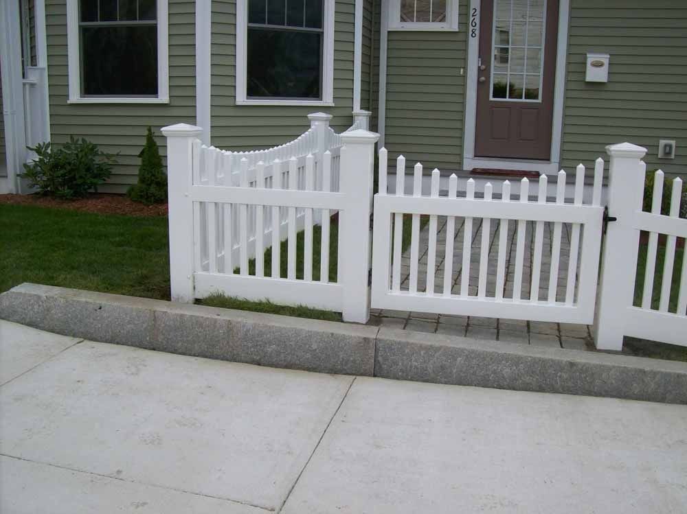 White picket fence in front of a house with green siding and a sidewalk.