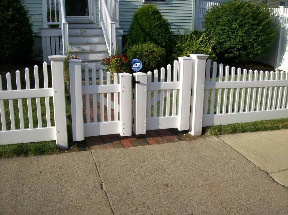 White picket fence and gate in front of a house with bushes and a sidewalk
