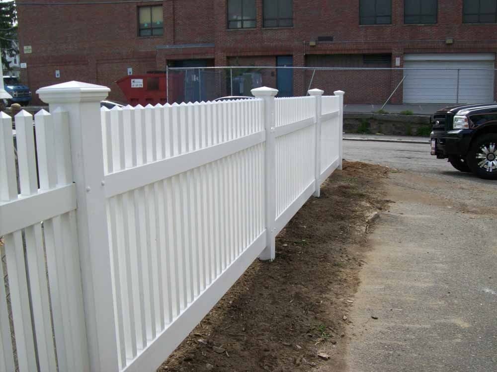 White vinyl fence along a dirt strip beside a driveway, with brick apartments in the background.
