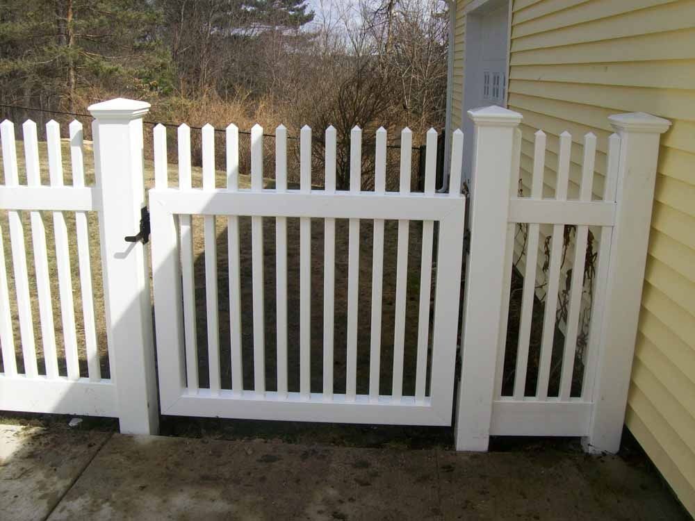 White picket fence gate beside a house, with a latch and side panel in a sunny yard.