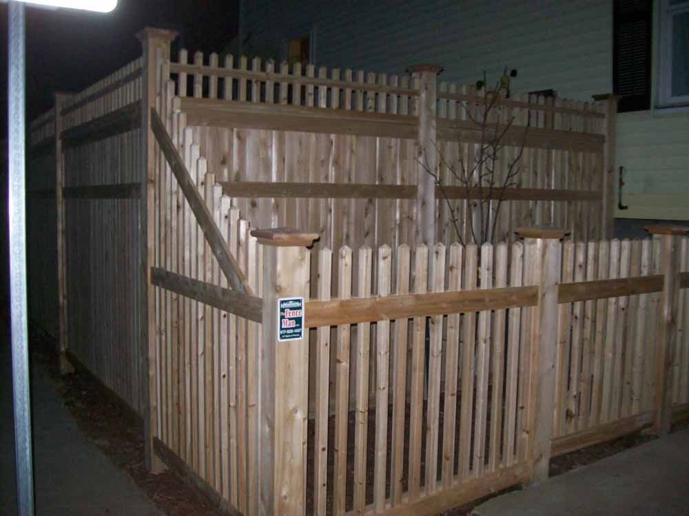 Wooden fenced enclosure with a gated entry, lit at night on a sidewalk.