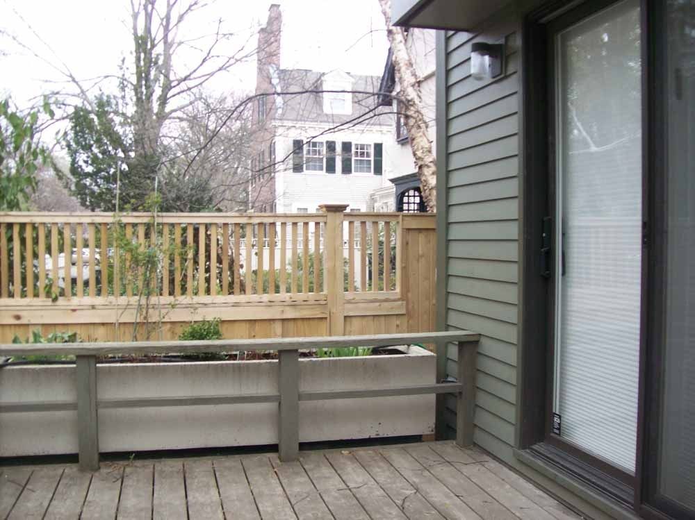 Wooden deck with bench seating beside a house, overlooking a fenced backyard and neighboring homes.