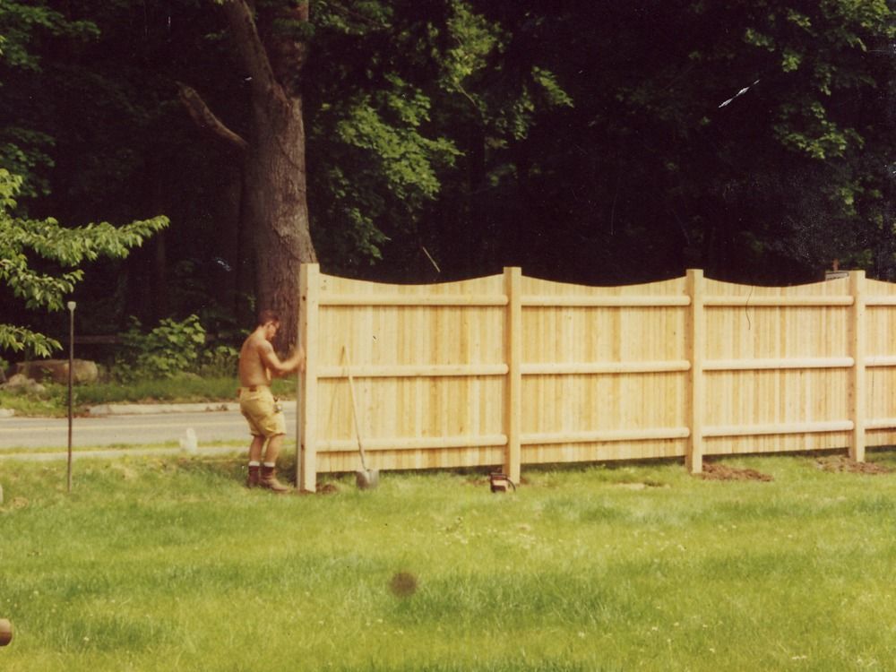 Person in tan clothing near a wooden fence in a grassy yard by trees