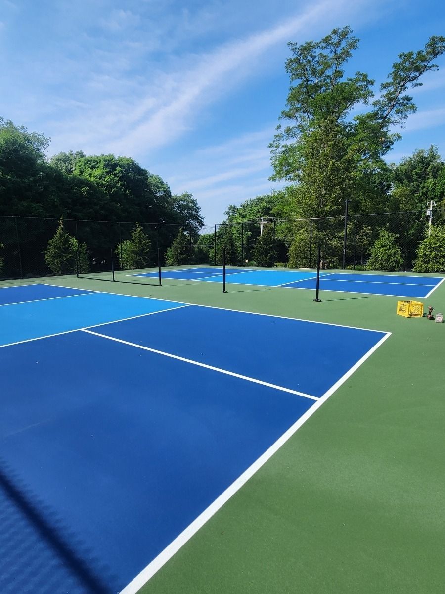 Outdoor tennis court with blue playing surface, green sideline area, and chain-link fence beside trees.