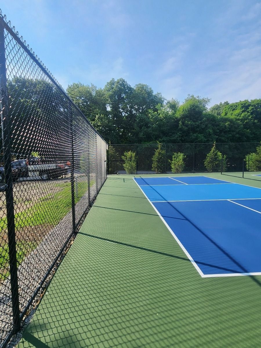 Blue outdoor tennis court beside a black fence, with trees and parked cars in the background.
