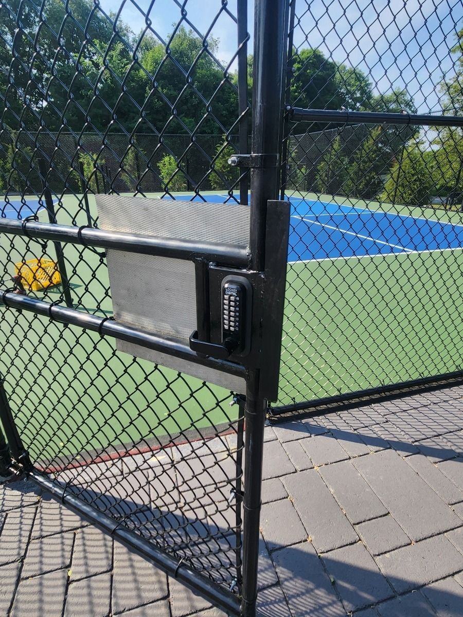 Black chain-link gate with keypad lock beside a tennis court and blue fence cover