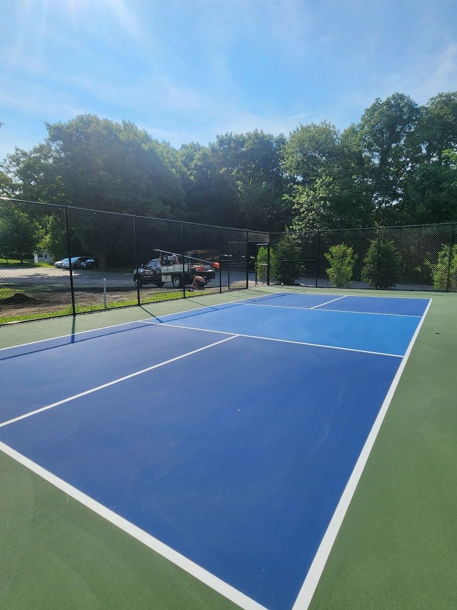 Blue outdoor pickleball court with net, green border, and trees in the background