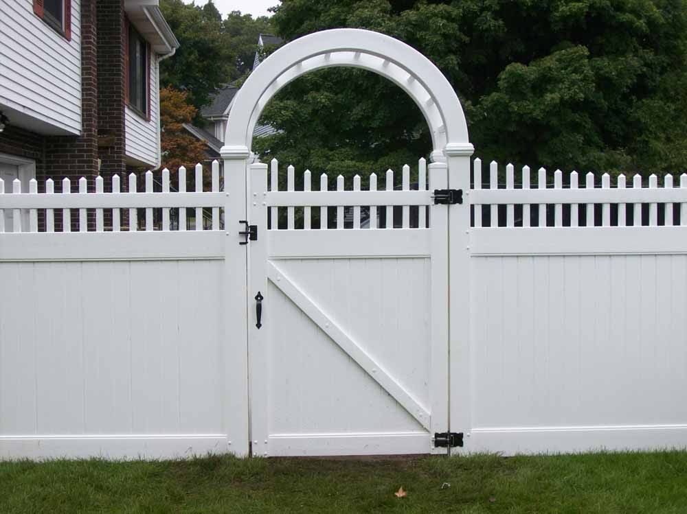 White picket fence with a small gate and arched arbor in front of a house and trees