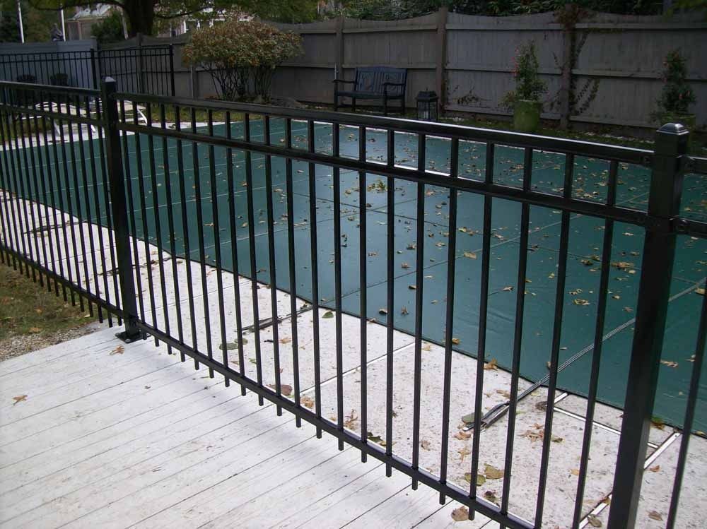 Black metal fence beside an empty outdoor swimming pool with a white deck and greenish water