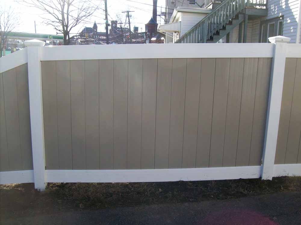 White-framed tan wooden fence panel along a sidewalk, with houses and trees visible above it.