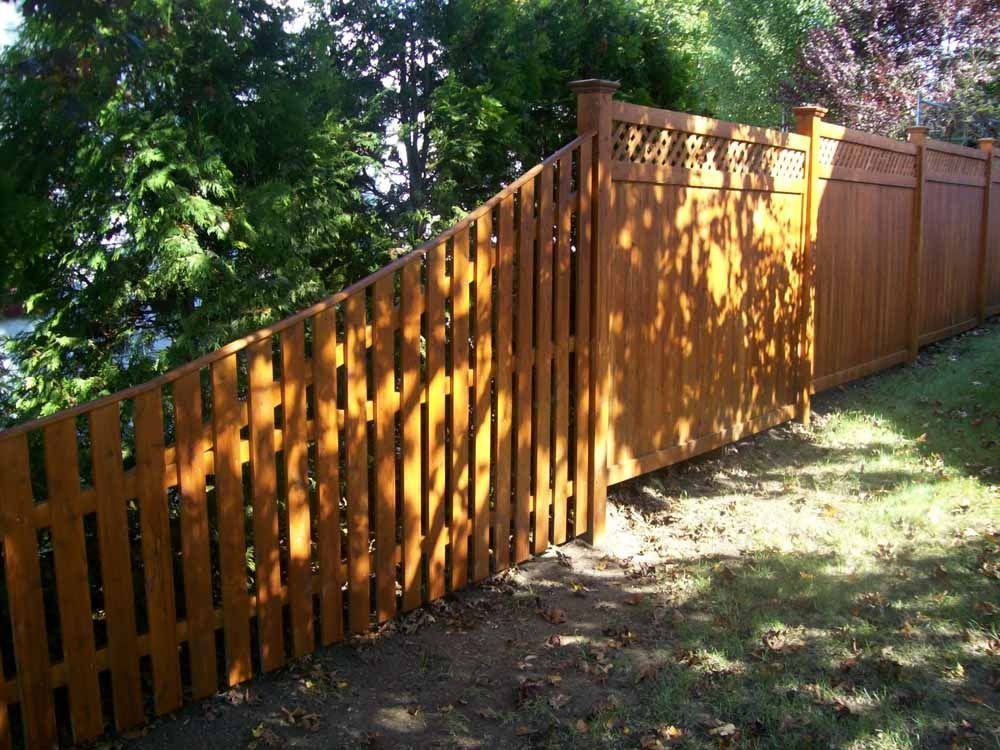 Wooden fence with a stepped slope, stained orange-brown, along a grassy yard with trees in sunlight