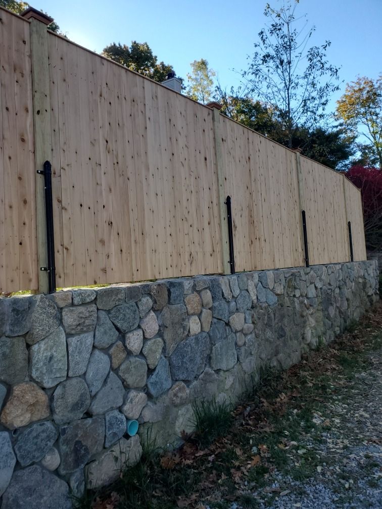 Stone retaining wall below a tall wooden fence on a sloped, wooded hillside.