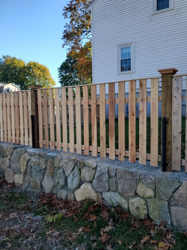 Wooden fence atop a stone retaining wall beside a white house in autumn.
