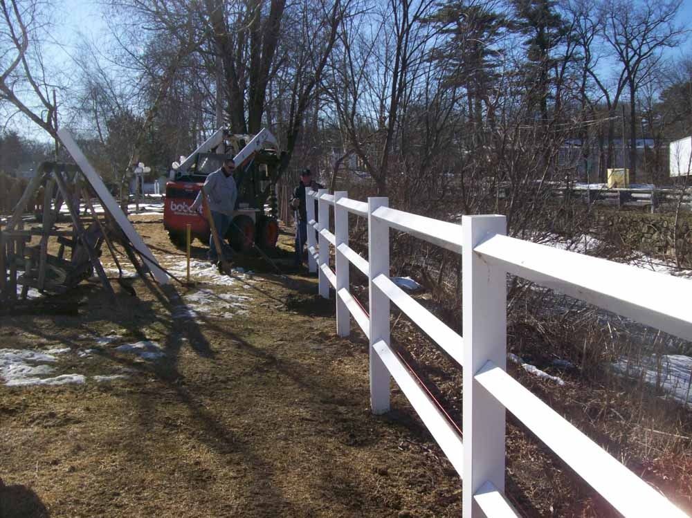 White fence beside a grassy yard with trees and a worker using a tractor or lift near a red truck