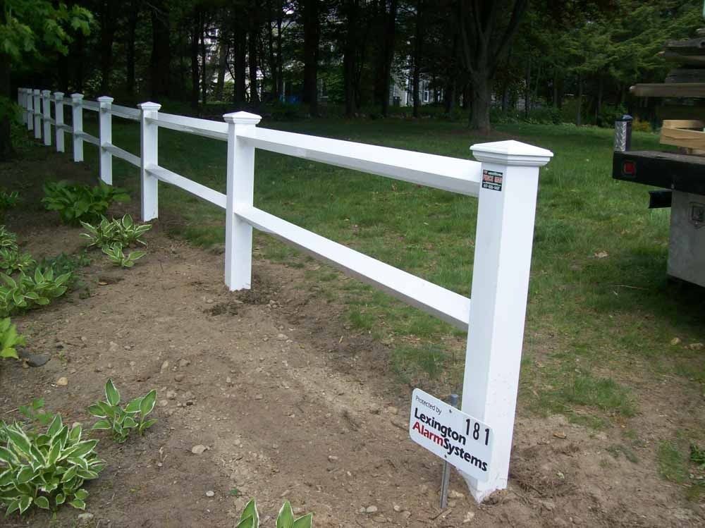 White wooden fence with a slanted rail along a dirt path beside trees and a parked vehicle.