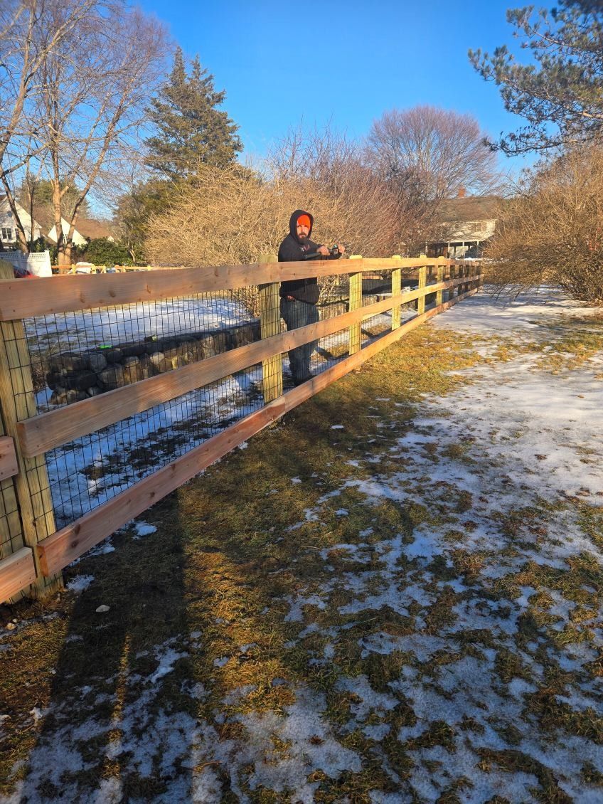 Person in a red jacket leaning on a wooden fence beside a snowy creek on a sunny winter day